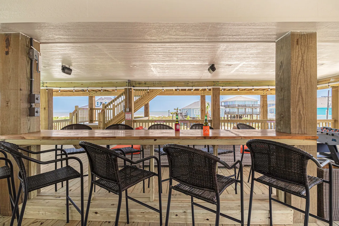 Covered outdoor patio area featuring a long wooden bar table with chairs, looking out toward neighboring beach houses and water in the distance.