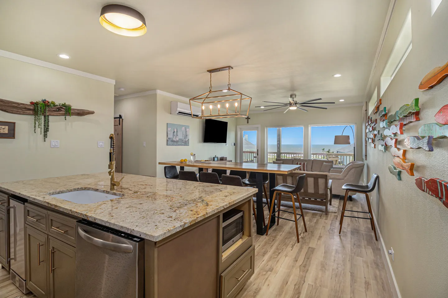 Open-concept living space with a kitchen island in the foreground, dining table, and living area overlooking water through windows.