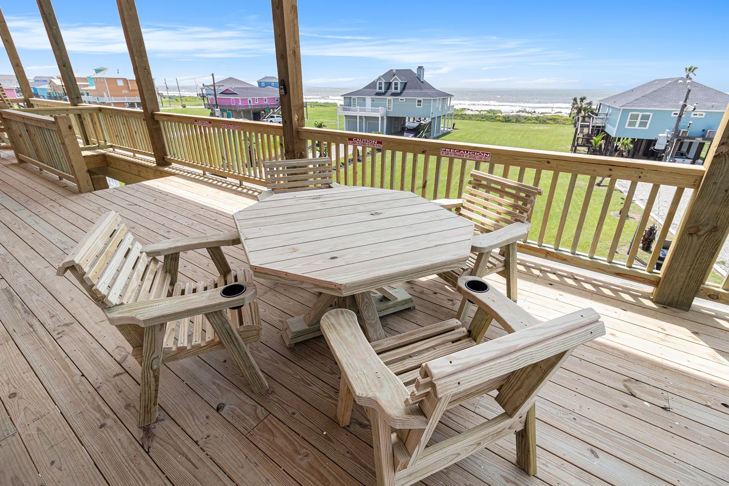 Elevated wooden deck with an octagonal wooden table, matching armchairs, and views of neighboring beach houses and the ocean.