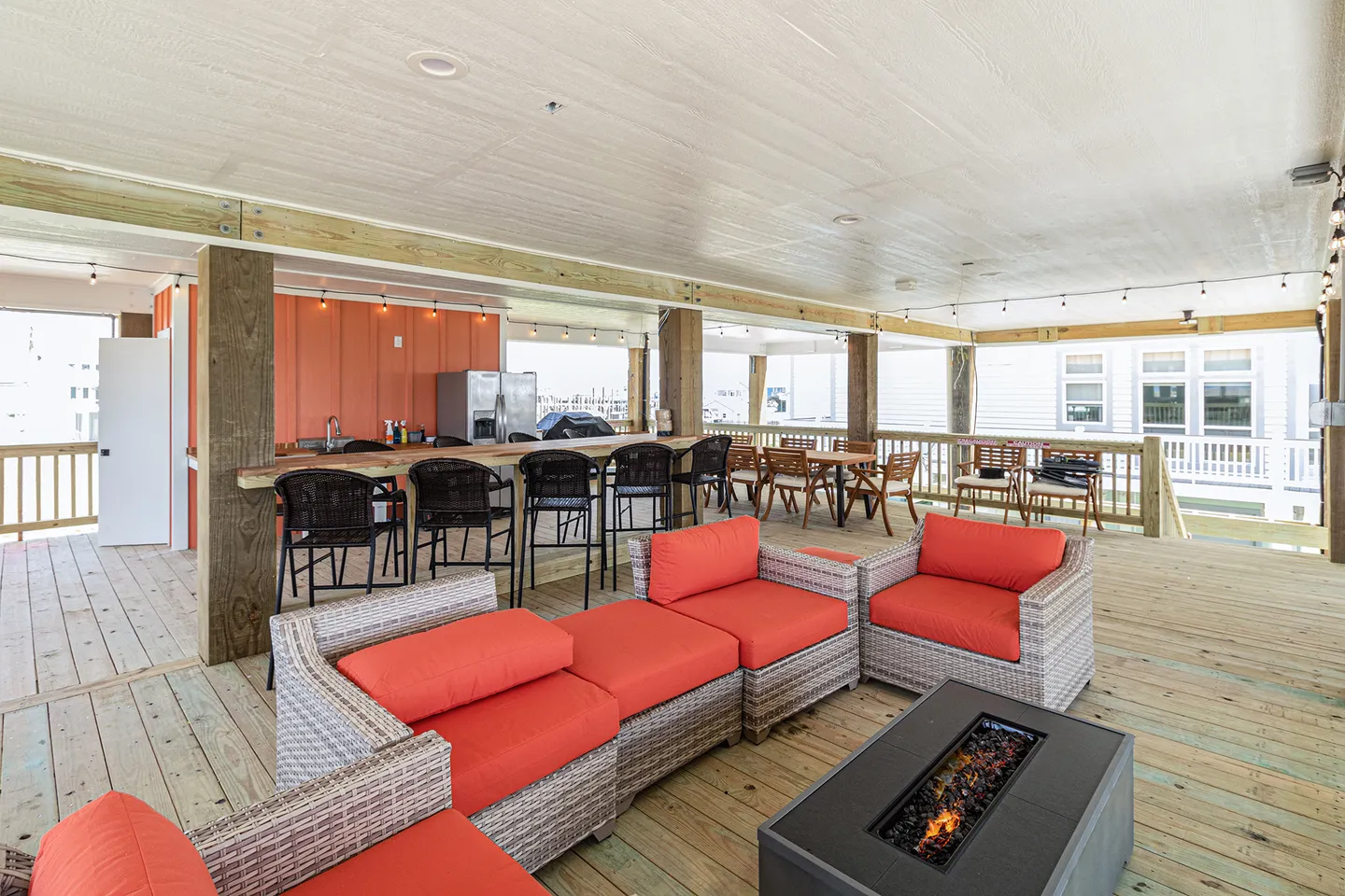 Covered outdoor deck with wicker seating, coral cushions, a fire pit table, bar area, and dining tables.