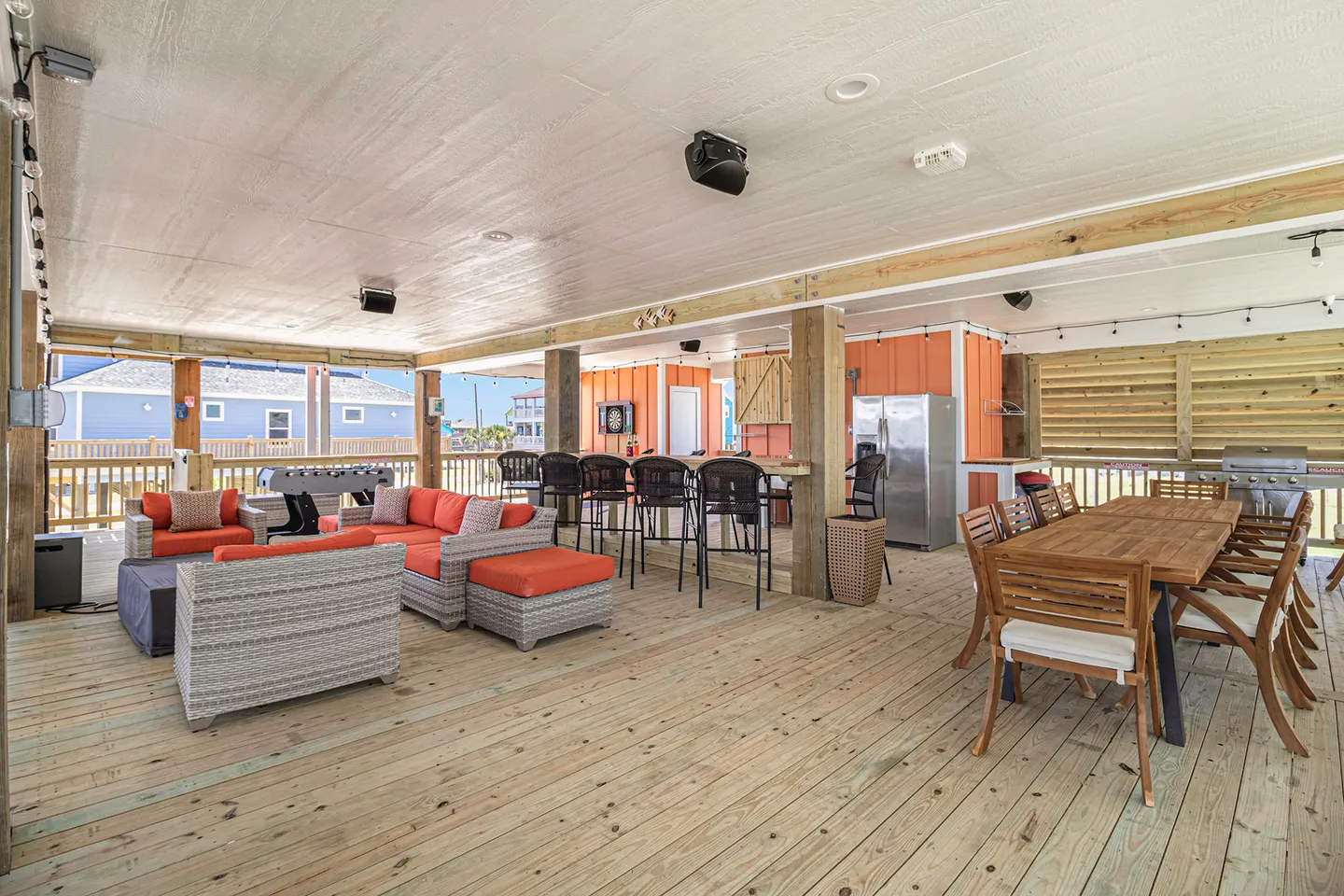 Spacious covered outdoor deck featuring wicker lounge seating with orange cushions, a wooden dining table, a bar counter with stools, and a foosball table.