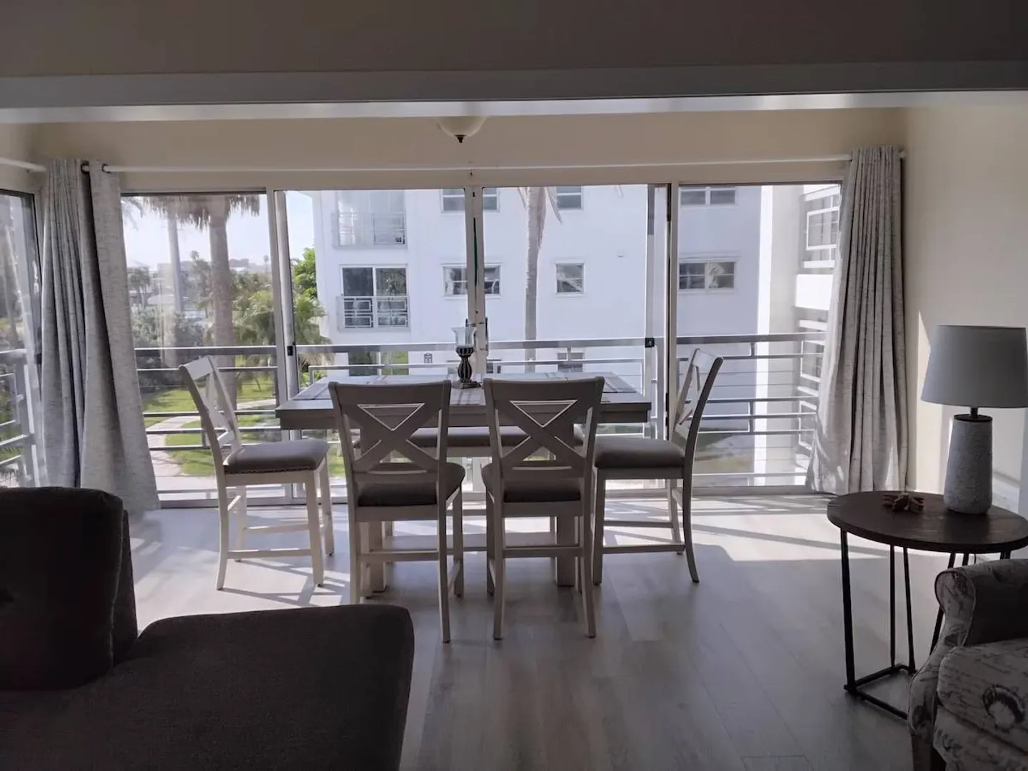 Dining area with a wooden table and high-back chairs positioned in front of sliding glass doors looking out toward palm trees and buildings.