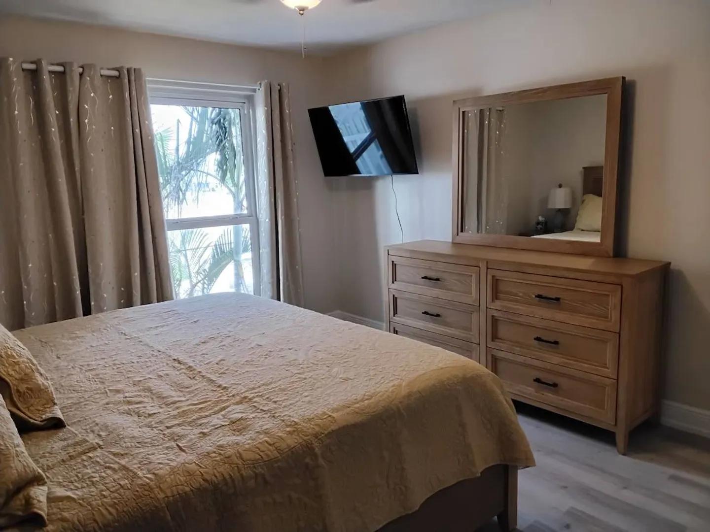 Bedroom with a bed made with a beige quilted spread, wooden dresser with a mirror, wall-mounted television, and a window with curtains.