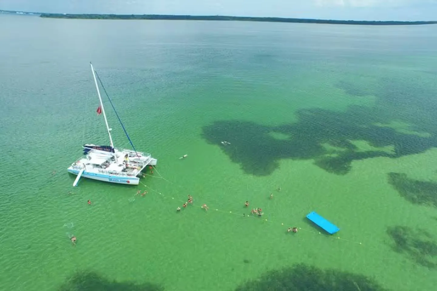 Aerial view of a catamaran anchored in clear turquoise and green shallow water with swimmers and a floating blue mat.