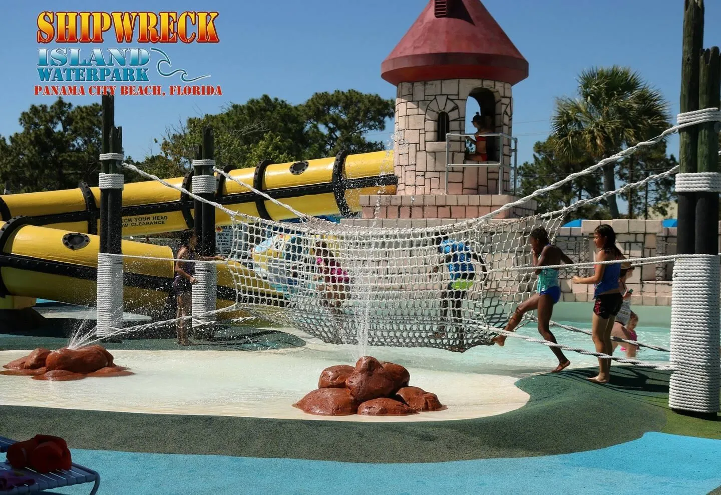 Outdoor waterpark play area featuring a rope bridge, splash fountain, small stone-style tower, and enclosed yellow water slide.