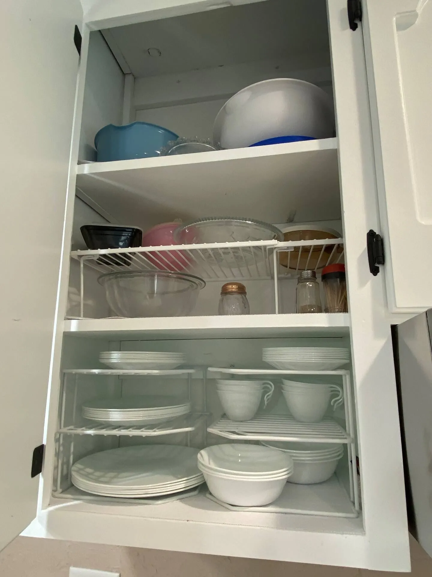 Open white kitchen cabinet stocked with white dishes, bowls, cups, and assorted mixing bowls on tiered wire organizers.