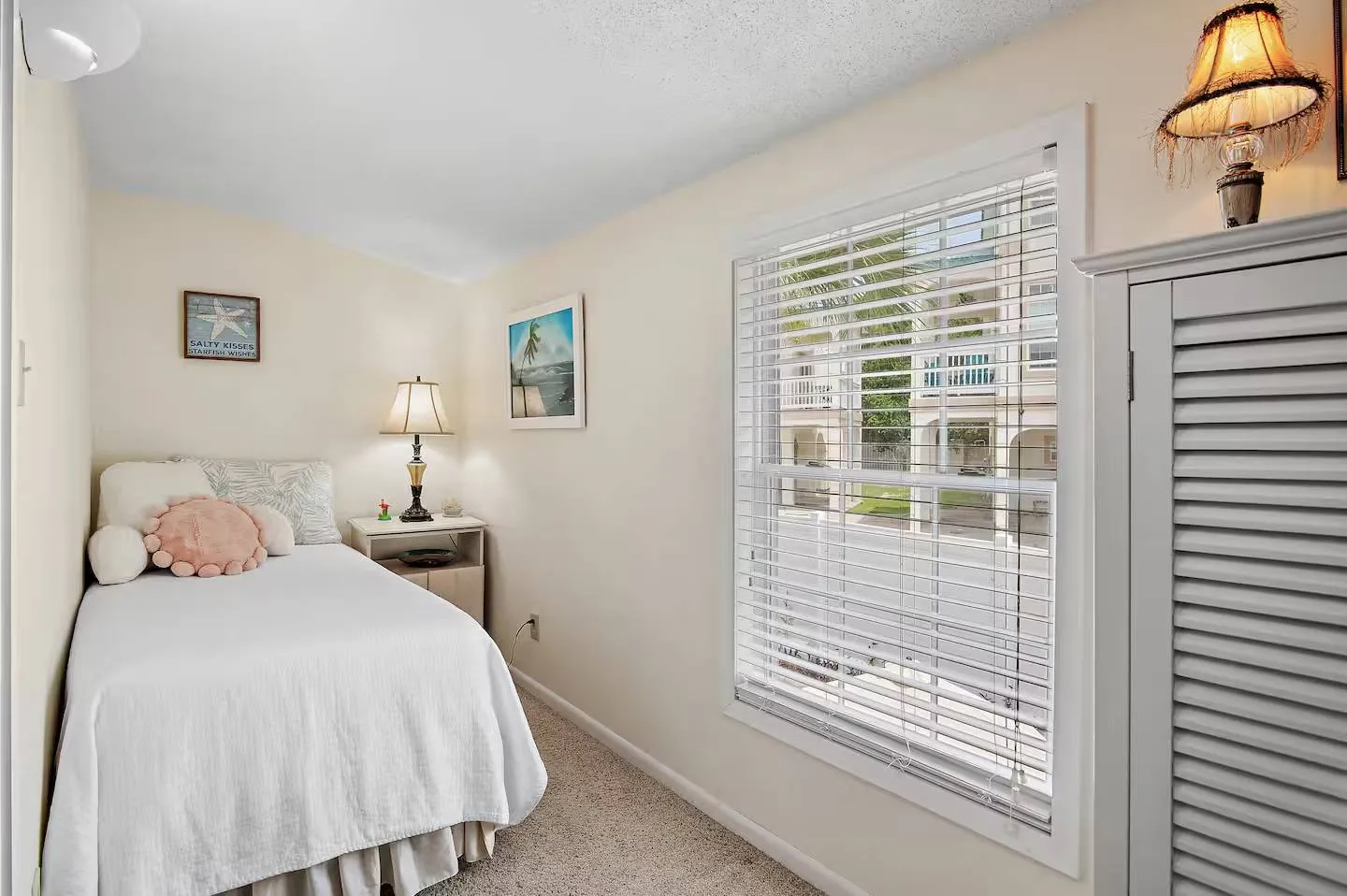 Bedroom with a single bed, carpeted floor, bedside table, and window with white blinds.