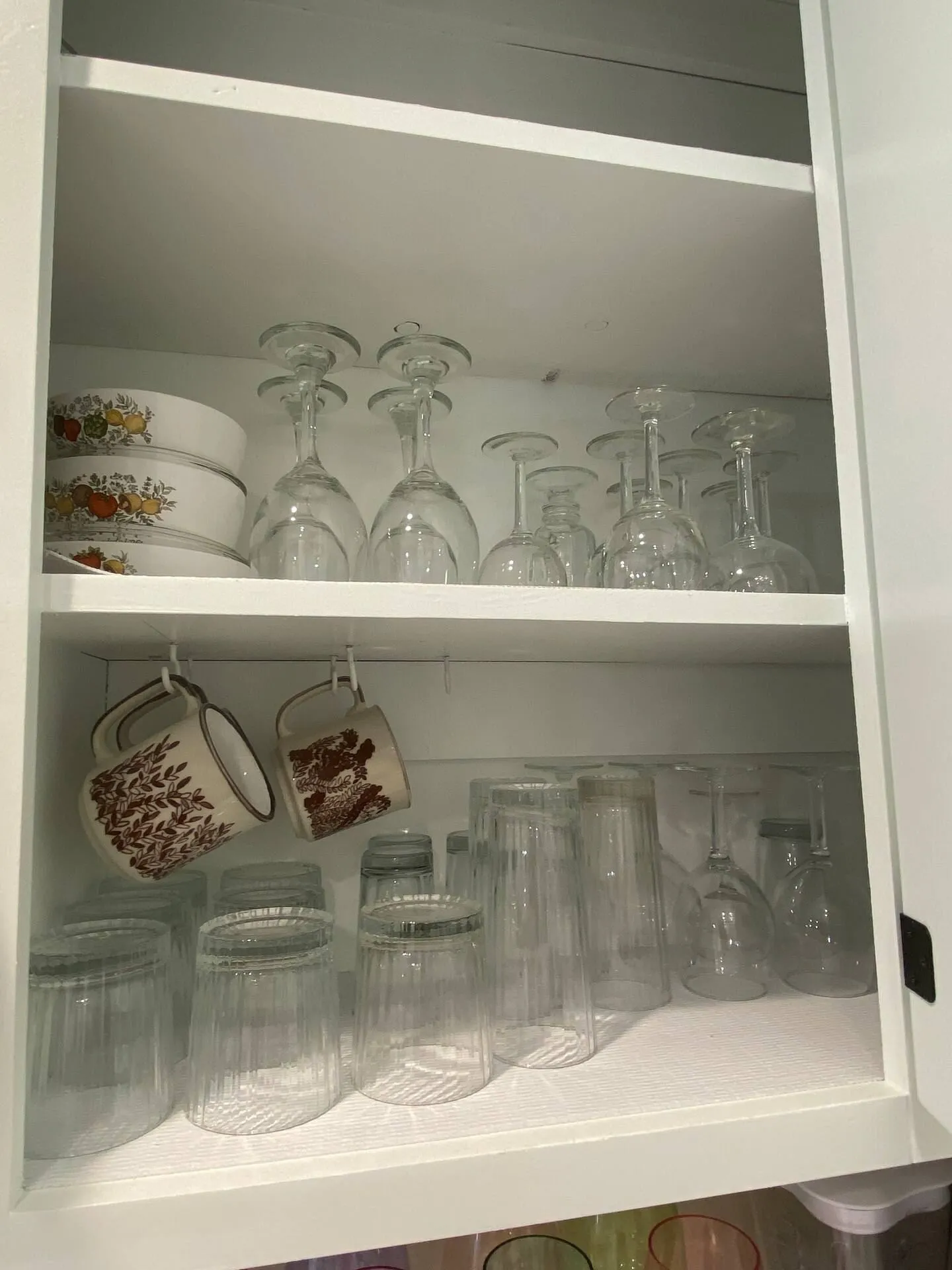 Open white kitchen cabinet shelves filled with drinking glasses, wine glasses, bowls, and hanging mugs.