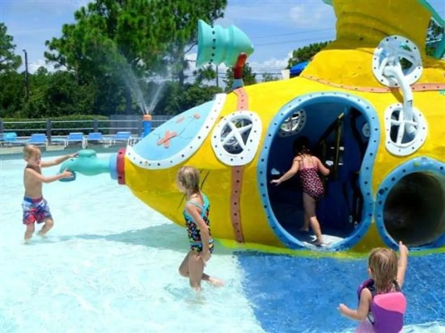 Children playing around a yellow submarine play structure in a shallow splash pool under bright sunlight.