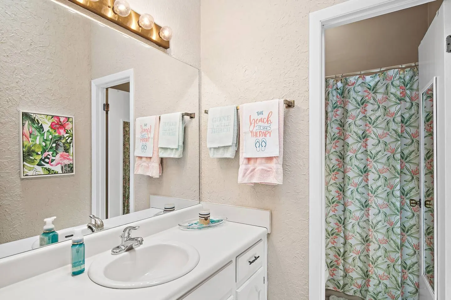 Bathroom vanity with a large mirror, globe light bar, white sink, and a view into the shower area with a floral curtain.