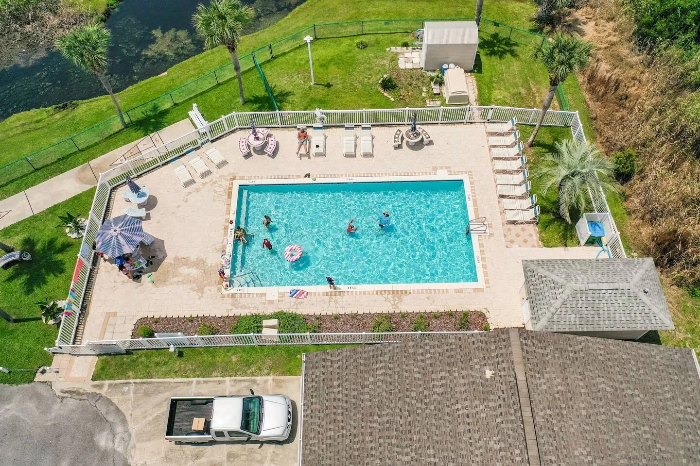 High-angle view of a fenced community swimming pool with swimmers, surrounded by lounge chairs, patio tables, and lawn.