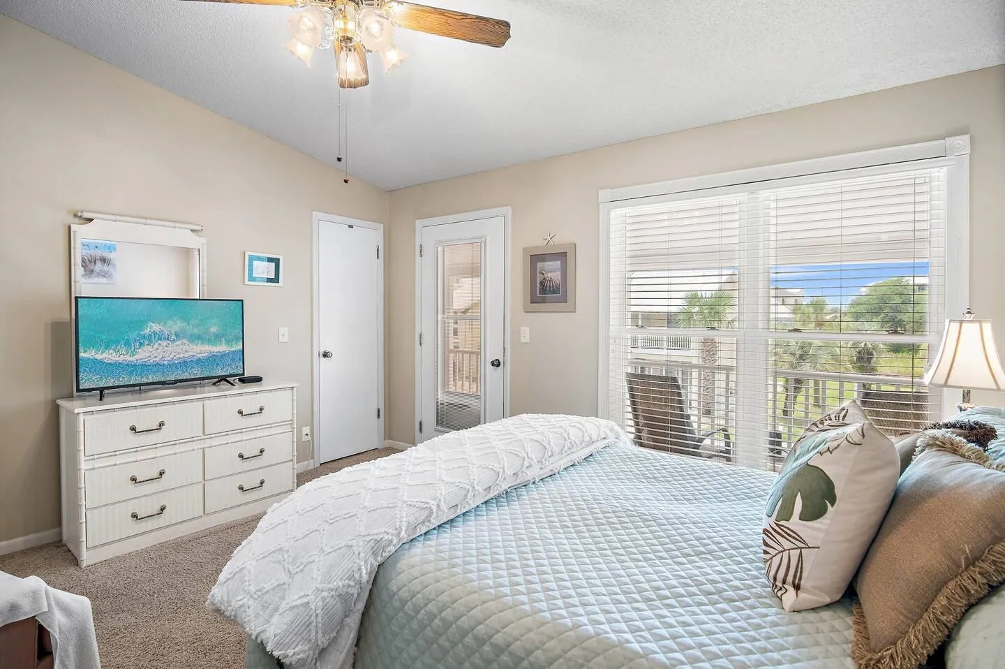 Bedroom with a made bed, dresser with a television, ceiling fan, and a glass door leading to a balcony.