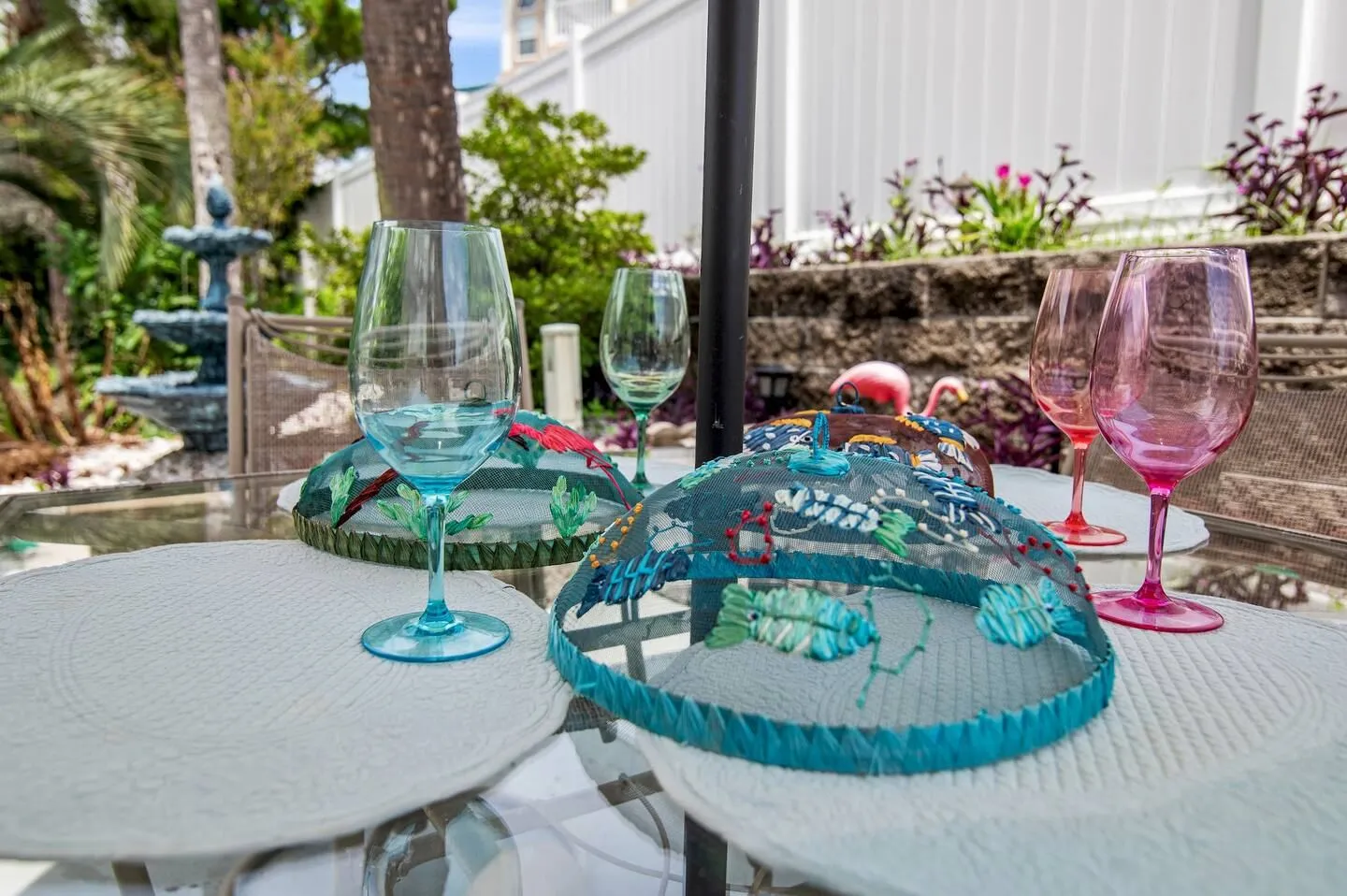 Close-up of an outdoor patio table set with colorful wine glasses, woven placemats, and decorative mesh food covers.