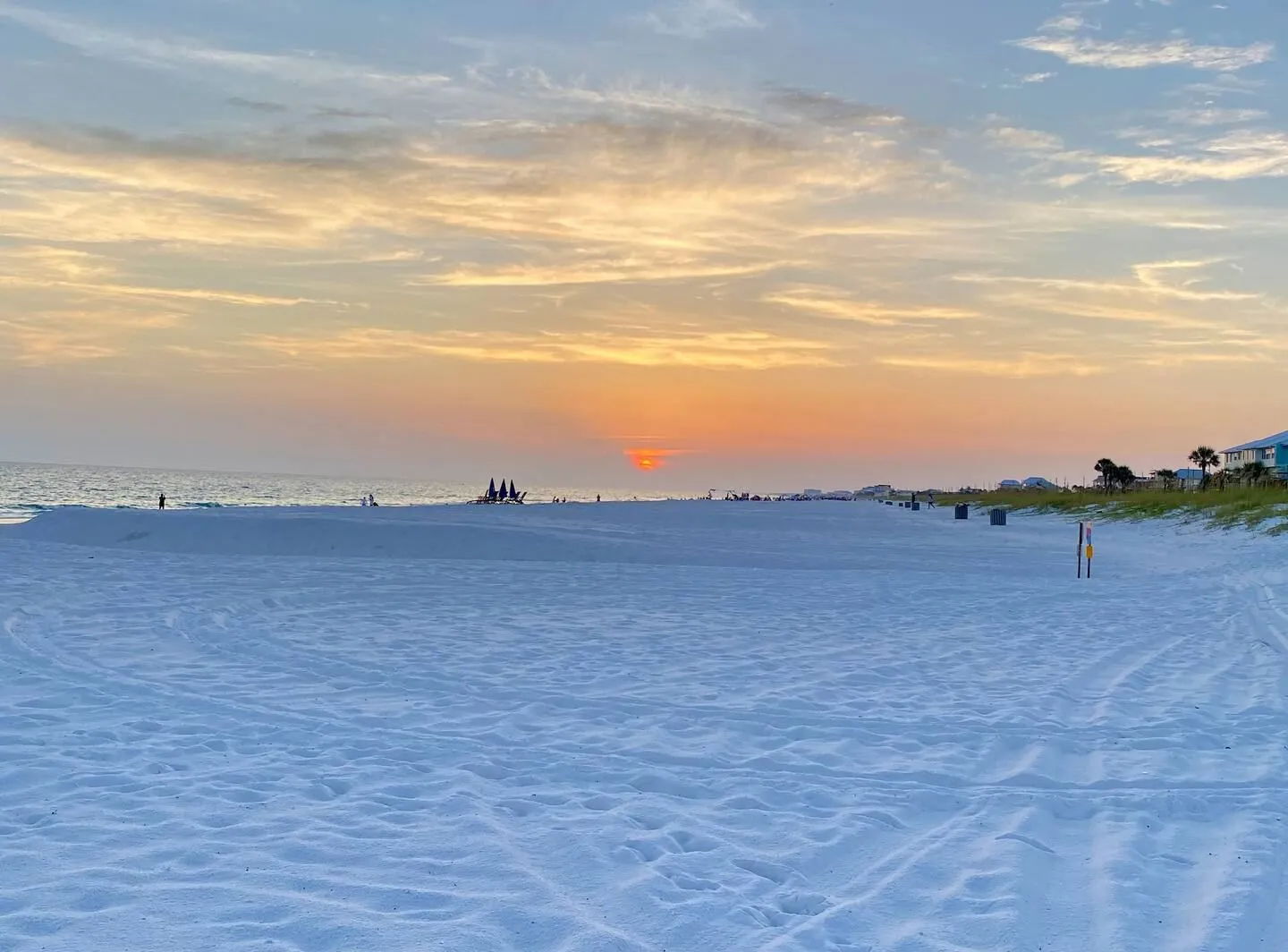 Wide view of a white sand beach at sunset with the sun dipping below clouds over the ocean.