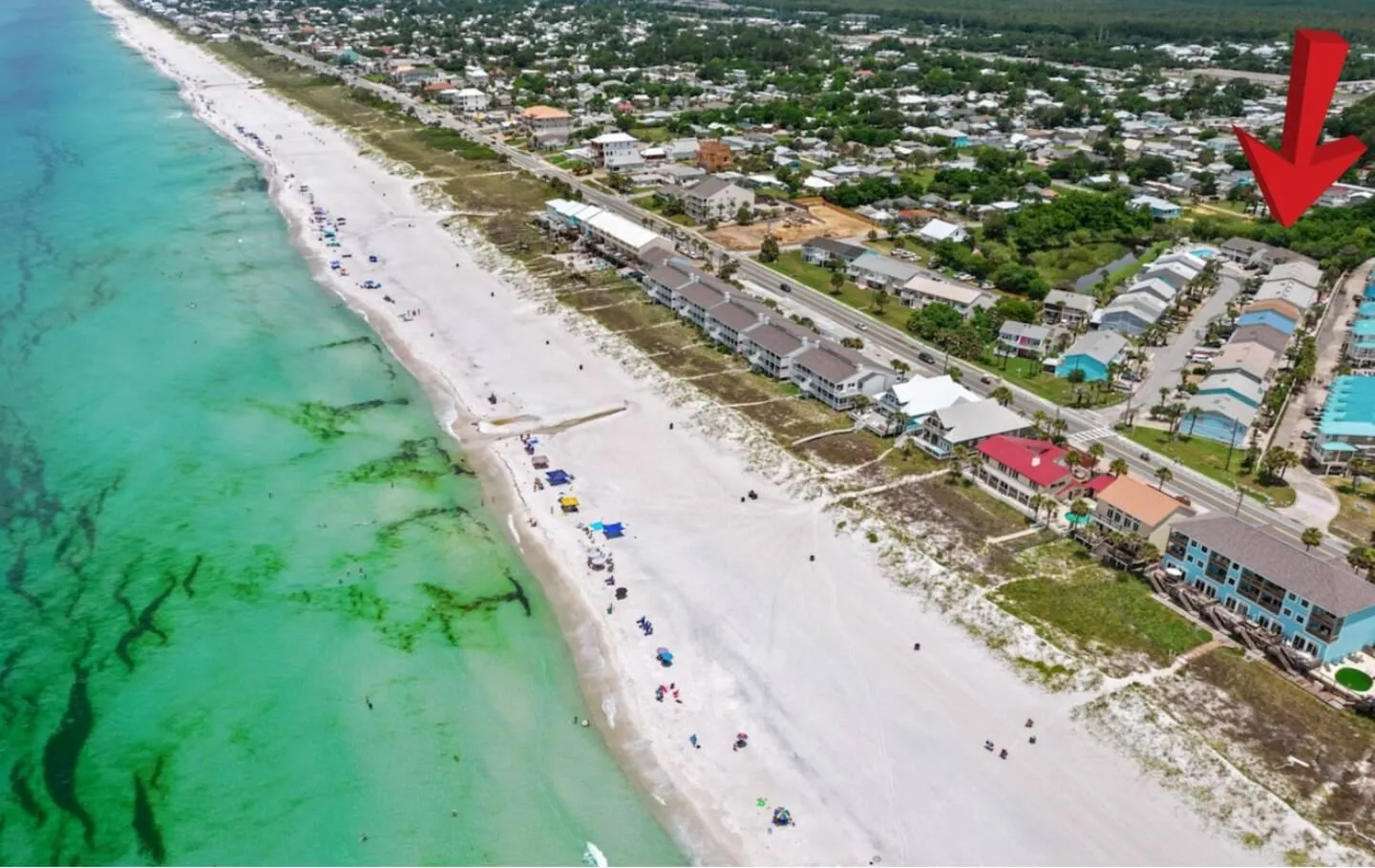 Aerial view of a white sandy beach with turquoise water and coastal homes marked with a red arrow.