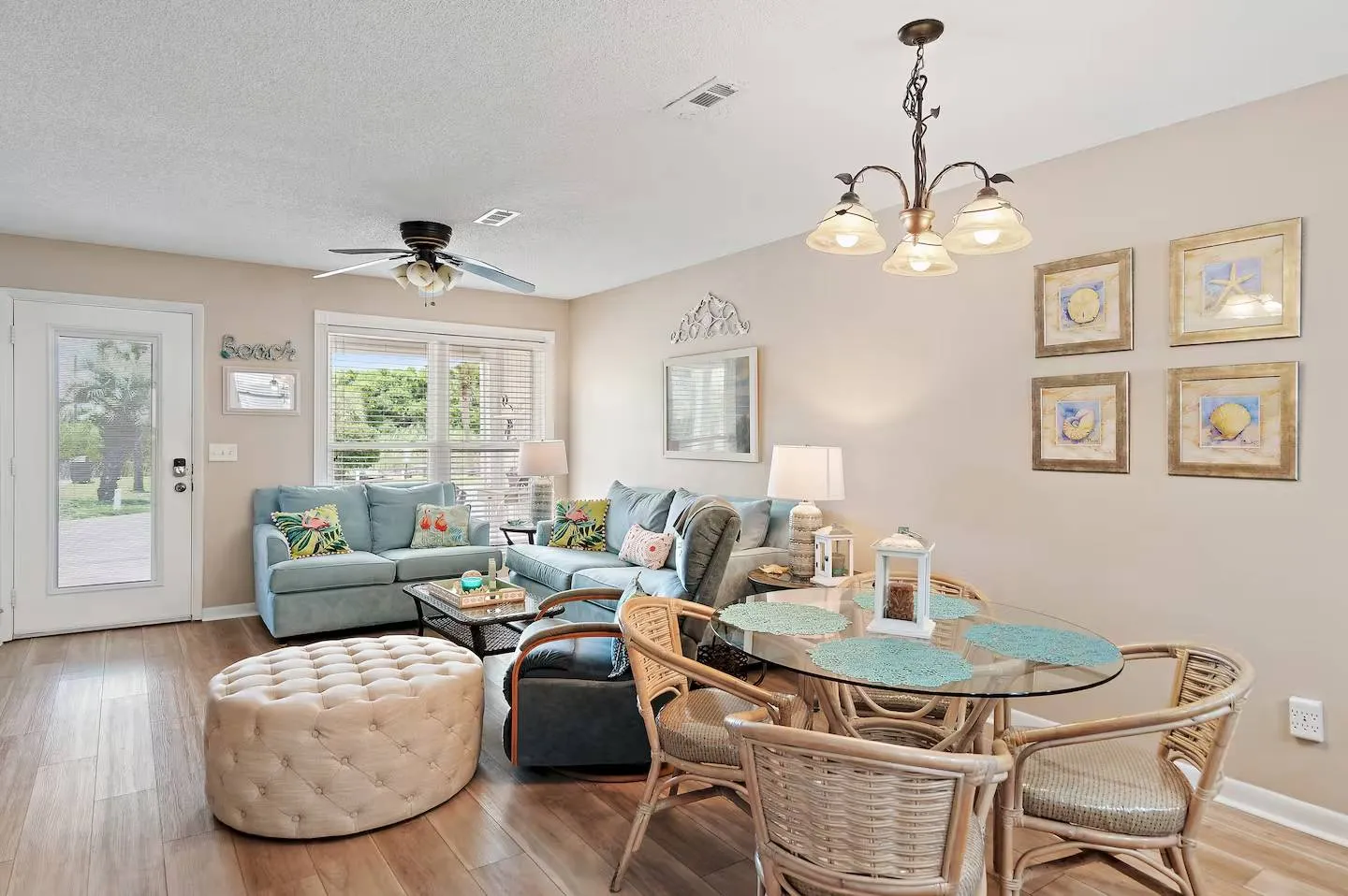 Open living and dining area with blue upholstered sofas, a tufted ottoman, and a glass-top dining table with wicker chairs.