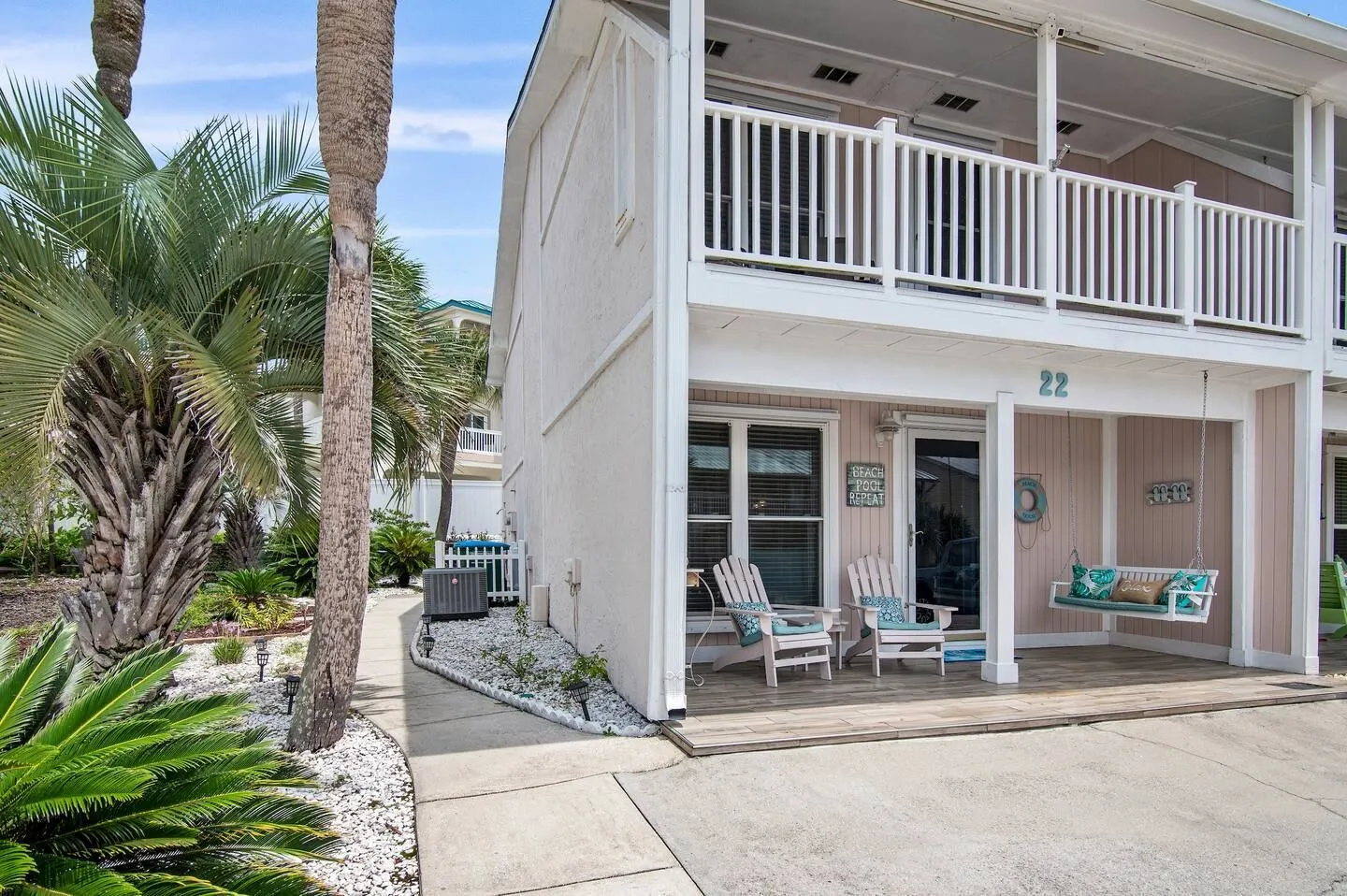 Exterior ground-floor patio with armchairs, porch swing, and palm trees in front of a townhouse unit.