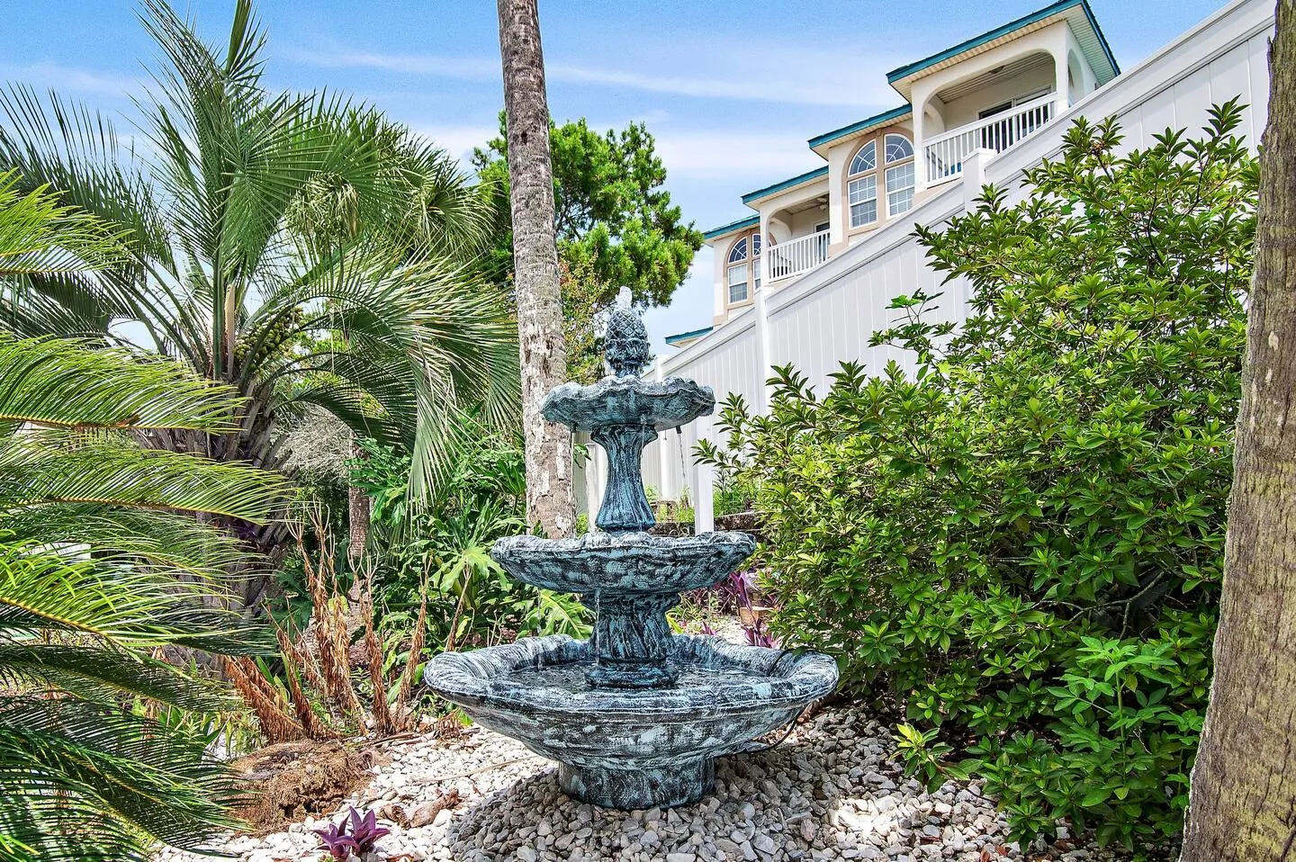 A tiered outdoor water fountain surrounded by rocks, palm trees, and tropical landscaping near a multistory house.
