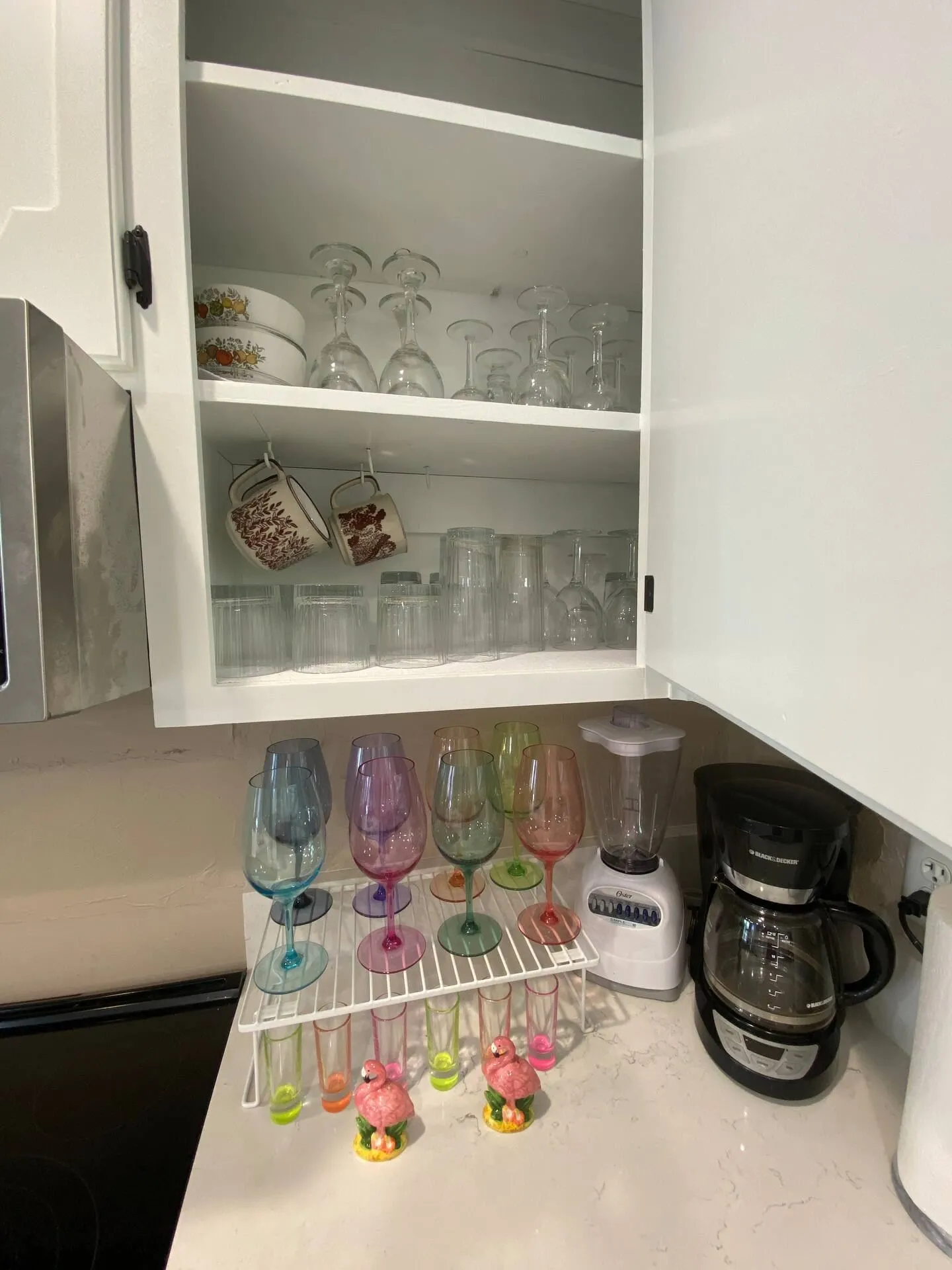 Open white kitchen cabinet stocked with glassware and mugs, with colorful wine glasses, a blender, and a coffee maker on the counter below.