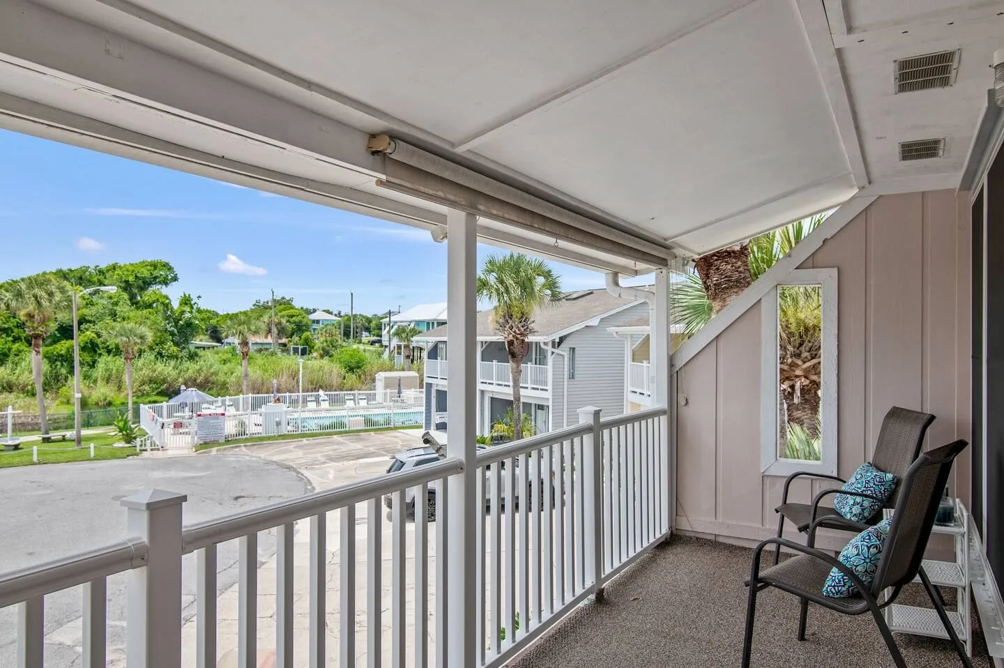 Covered balcony with white railing, armchairs, and a view of the community parking area and fenced pool.