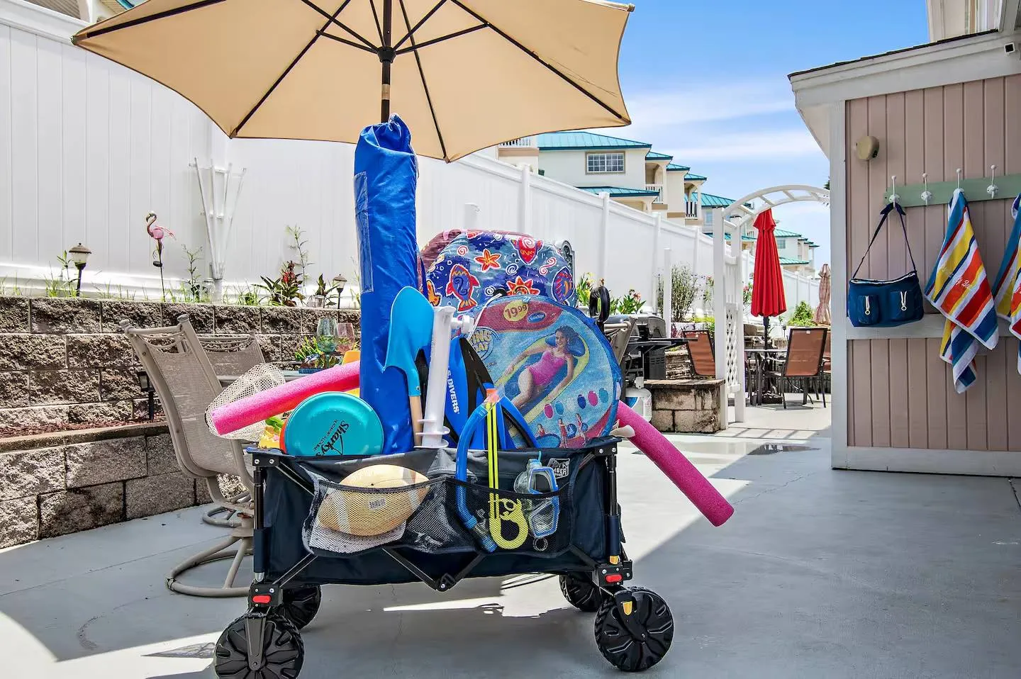 A wheeled beach cart loaded with pool noodles, snorkels, floats, and an umbrella on an outdoor patio.