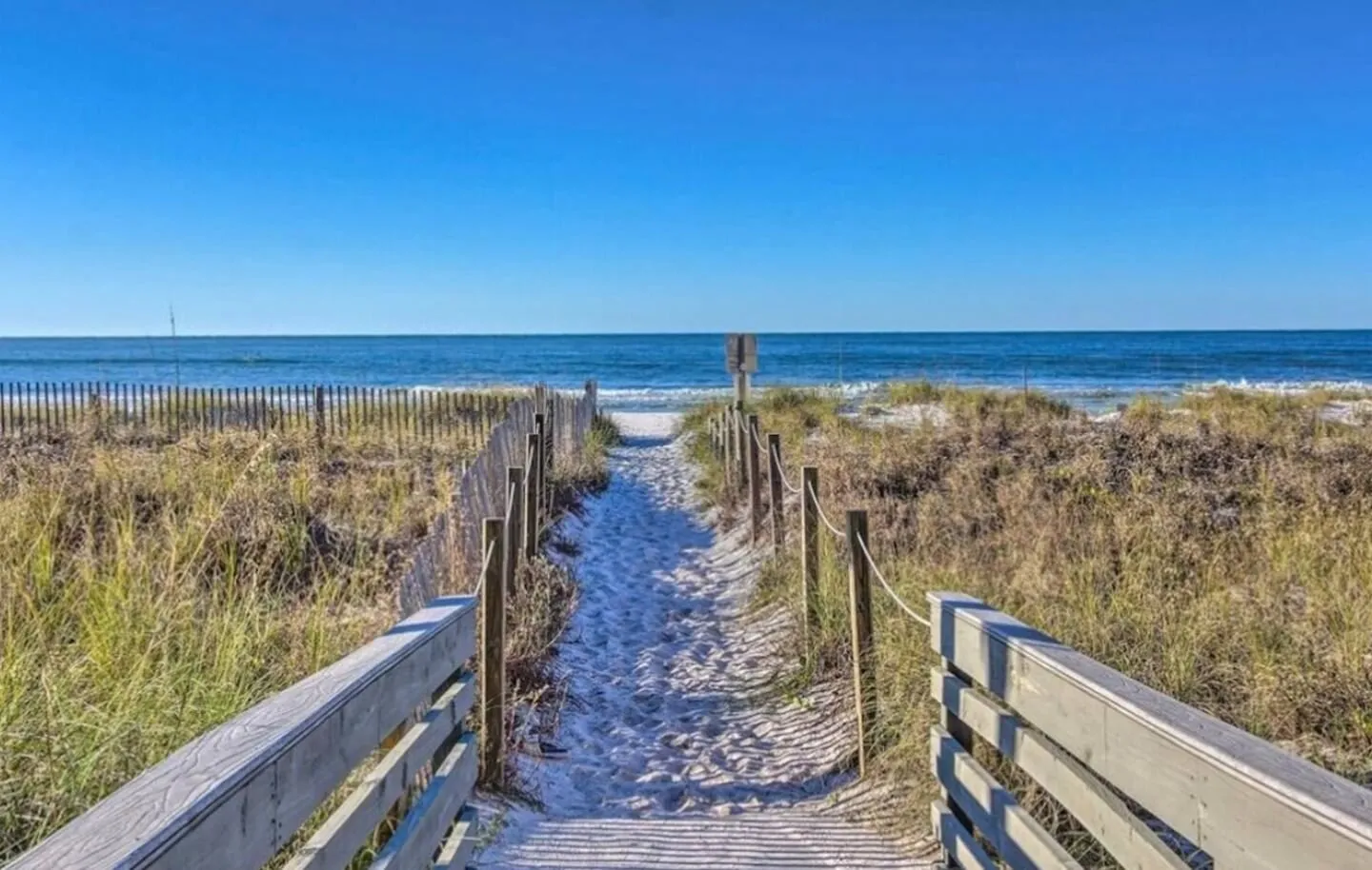 A sandy pathway with wooden railings and fencing leading through coastal dunes directly to the ocean.