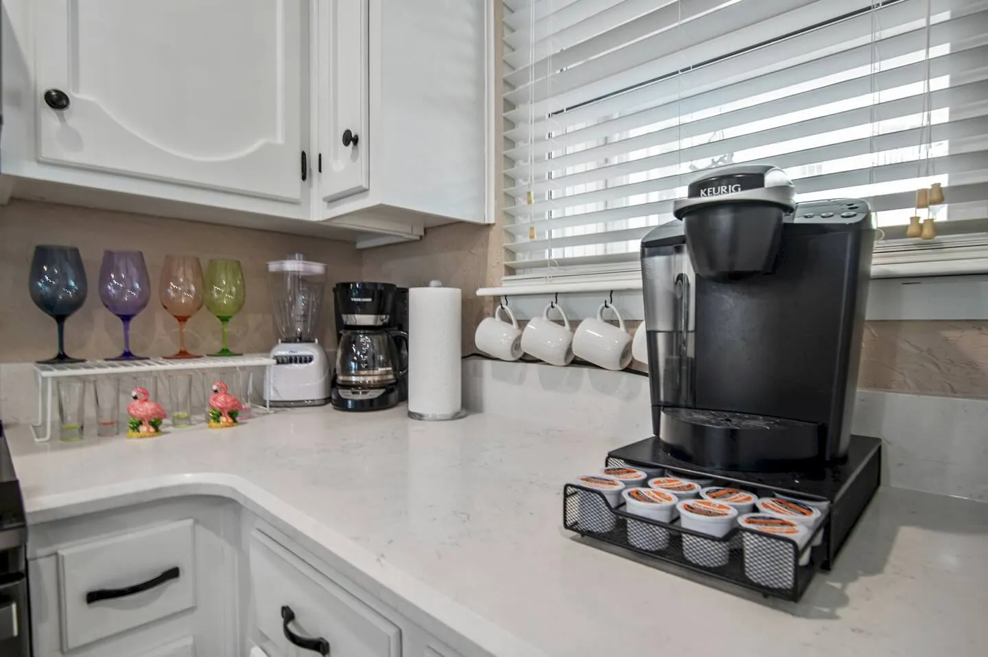 Kitchen countertop with a Keurig single-serve brewer, drip coffee maker, blender, glassware, and mugs hanging beneath a window with blinds.