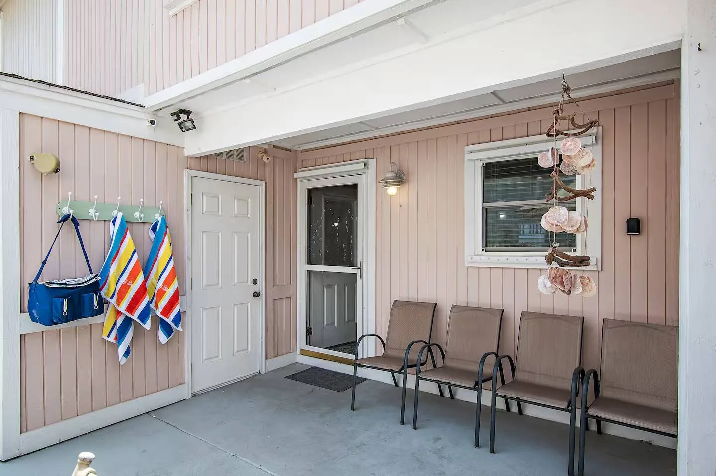 Covered patio entryway featuring light pink vertical siding, a row of chairs, and towels hanging on a wall rack.
