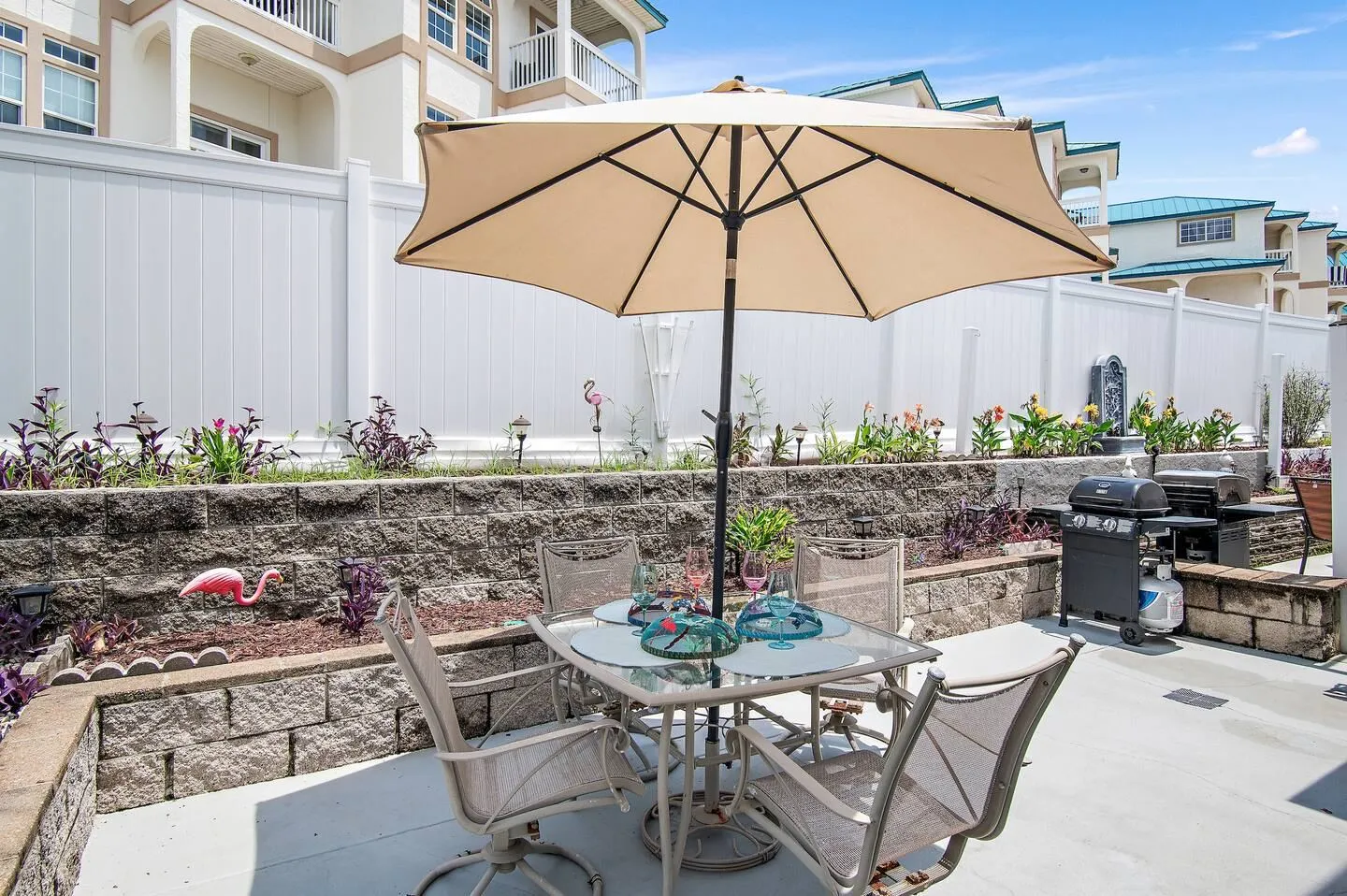 Outdoor patio with an umbrella-shaded dining table, chairs, and a barbecue grill next to retaining wall planters.