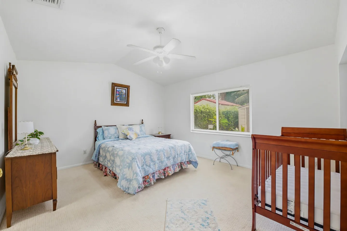 Bedroom with carpet, a made bed with floral bedding, a wooden crib, a dresser, and a window.