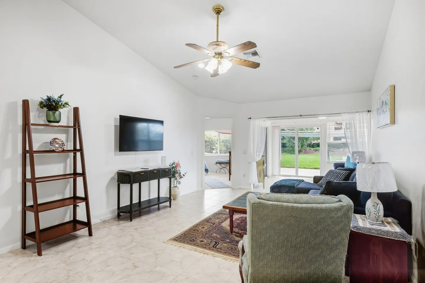 Living room with vaulted ceiling, ceiling fan, wall-mounted television, tiled floors, armchairs, and sliding glass doors to the backyard.