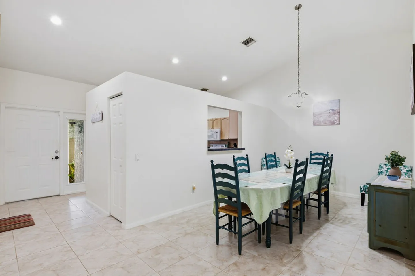 Dining area with tiled floors, a dining table surrounded by dark teal chairs, recessed lighting, and a view toward the front entryway and kitchen pass-through.