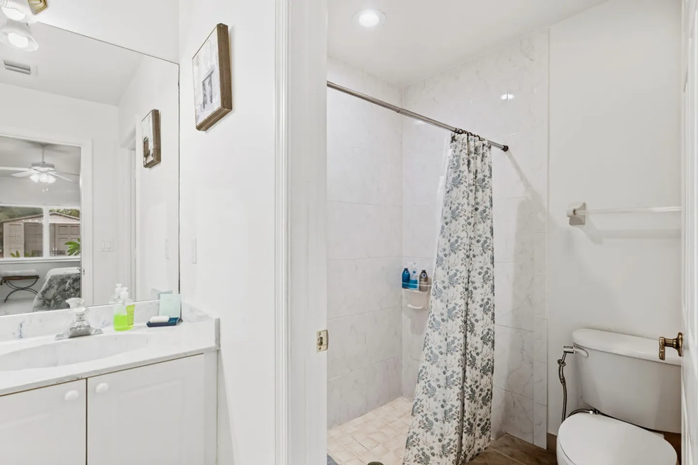 Bathroom showing a vanity sink on the left, an open tiled shower with a patterned curtain, and a toilet on the right.