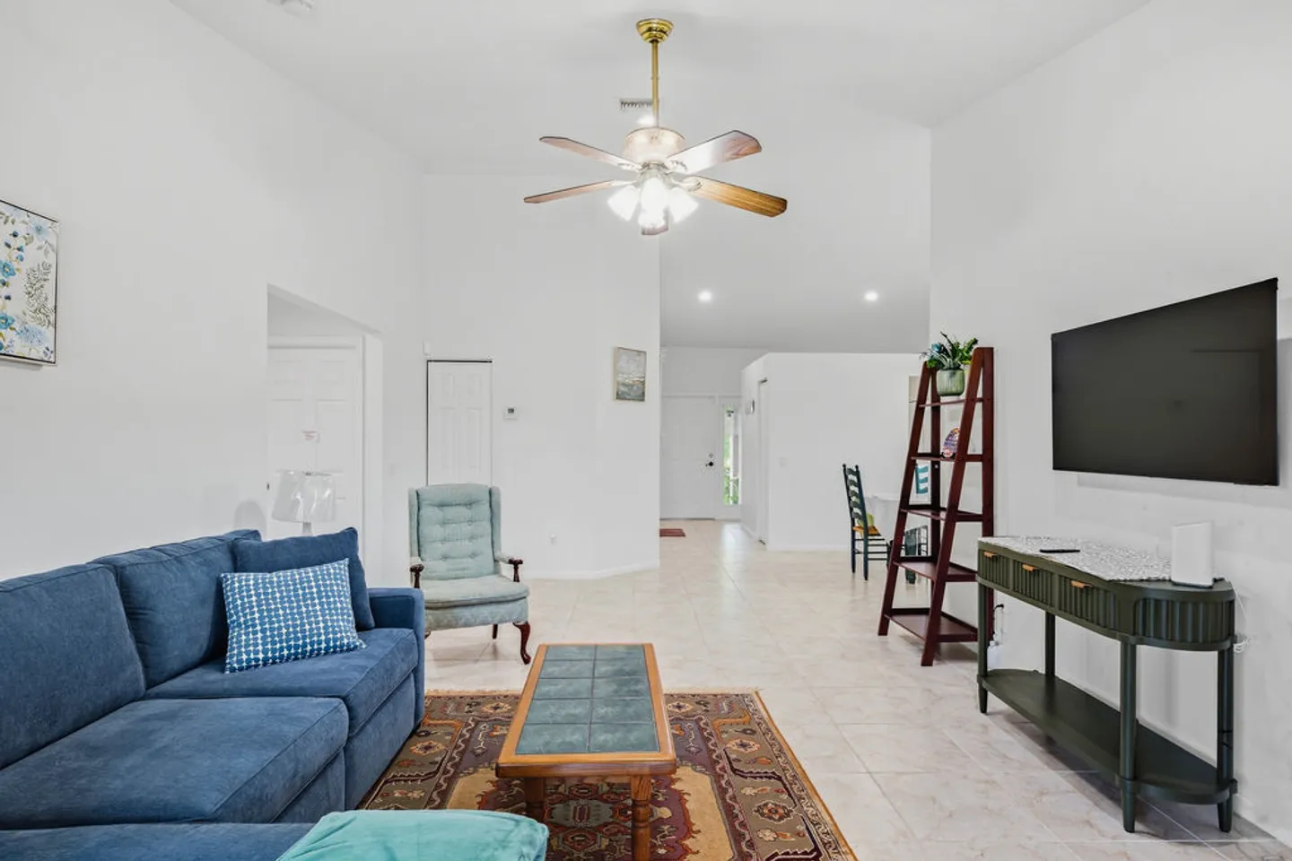 Living room with blue sofa, tiled floors, ceiling fan, wall-mounted TV, and an open layout view towards the hallway.