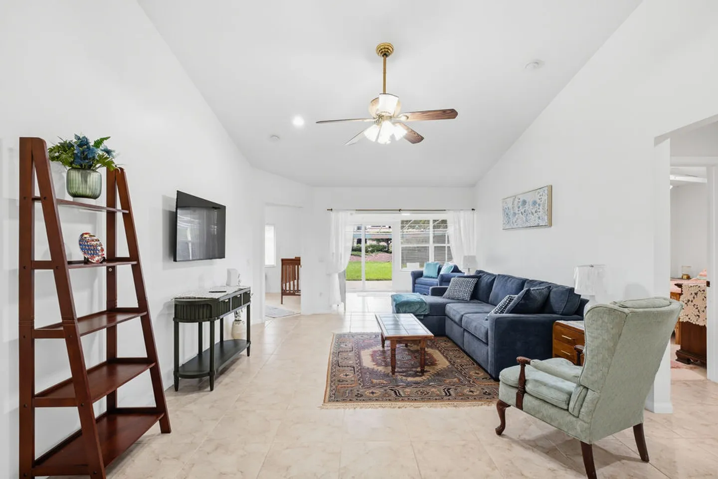 Bright living room with tiled flooring, vaulted ceiling, blue sectional sofa, armchair, and sliding glass doors leading to the yard.
