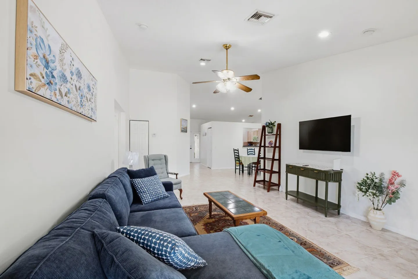 Living room with a large blue sectional sofa, a wall-mounted TV, a coffee table on a patterned rug, and a view into the adjoining dining and kitchen area.