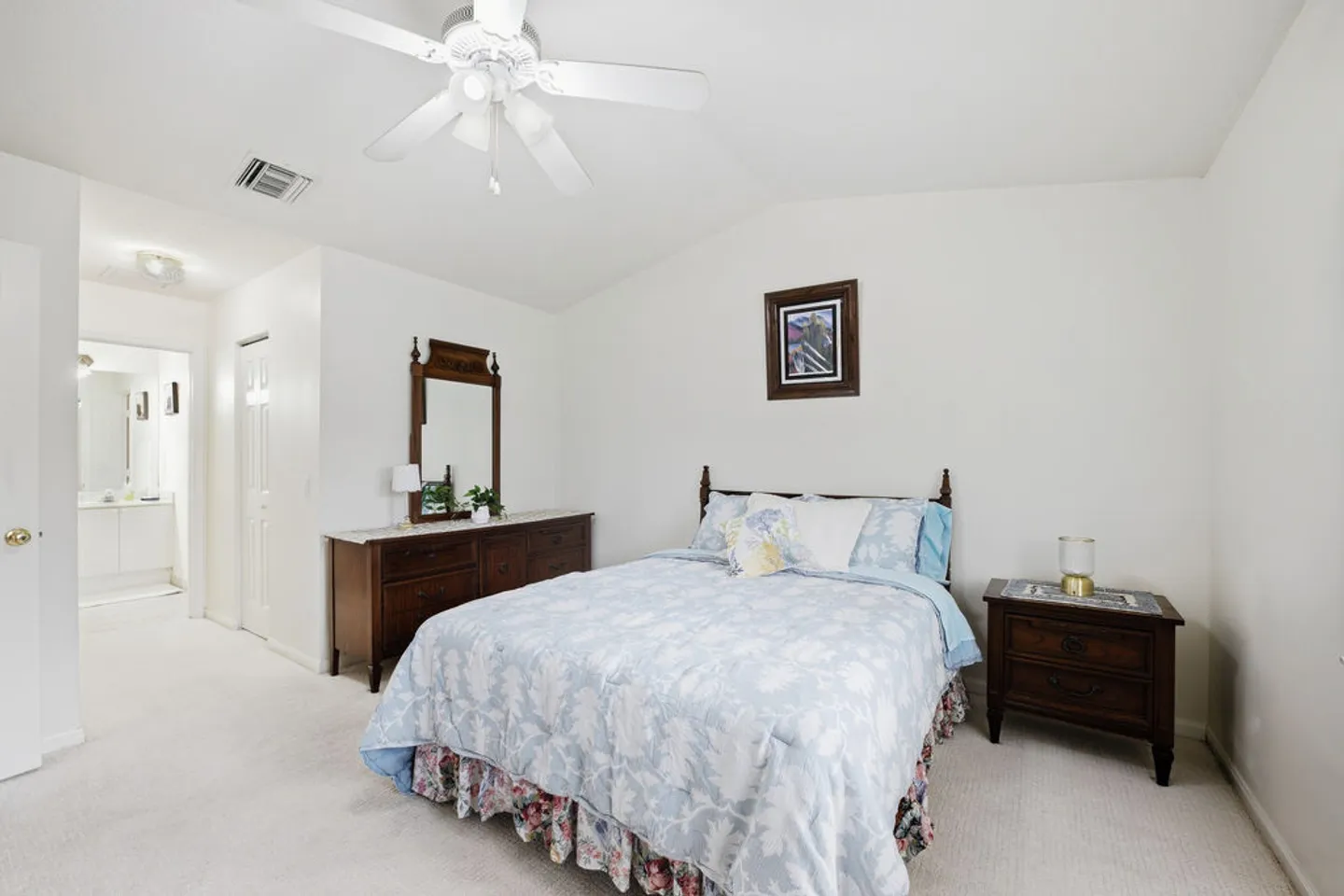 Bedroom with carpeted floors, a double bed with blue floral bedding, dark wood dresser with mirror, and attached bathroom.