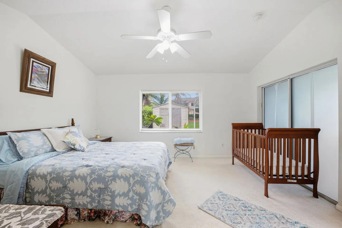 Bedroom with a patterned blue-and-white bed, a wooden crib, white carpet, a ceiling fan, and sliding glass doors.