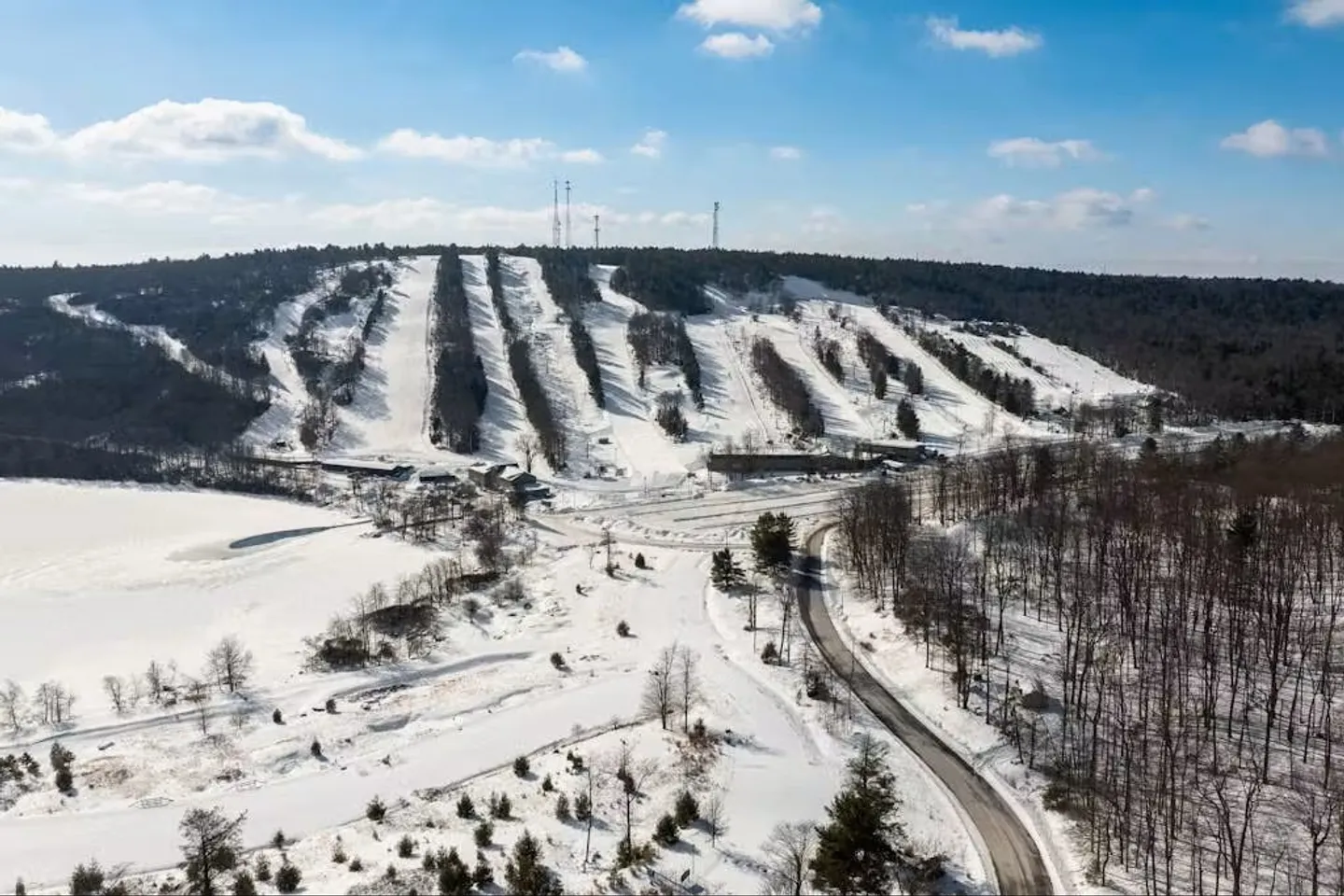 Aerial view of a snow-covered ski hill with cleared trails, a base area, and surrounding winter forest under a blue sky.