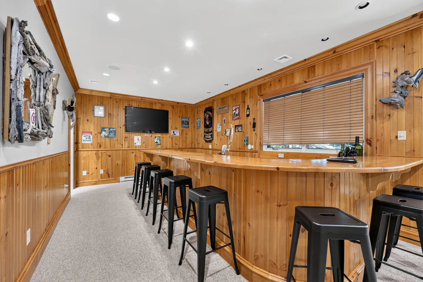 Finished basement bar area with wood-paneled walls, carpeted floor, long curved bar with black stools, and a wall-mounted TV.