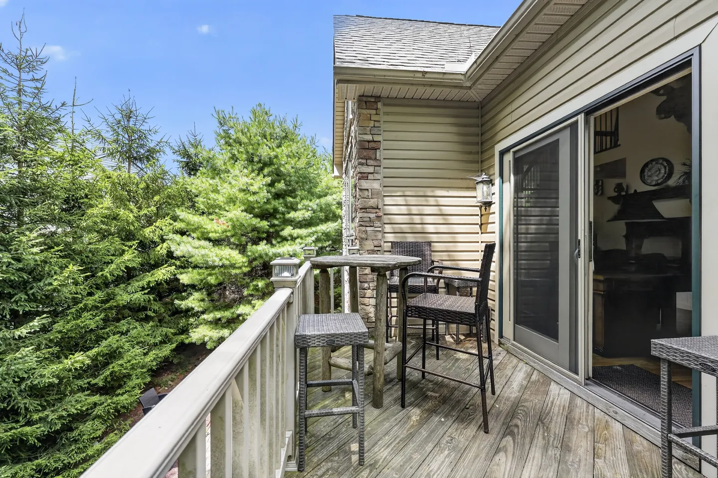 Outdoor wood deck with a small high-top table, wicker barstools, and sliding screen doors next to pine trees.