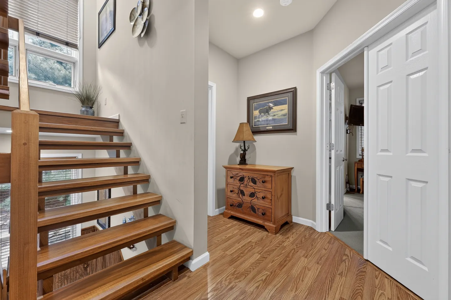 Open-riser wooden staircase and hallway with wood-look flooring, a dresser with a lamp, and an open doorway.