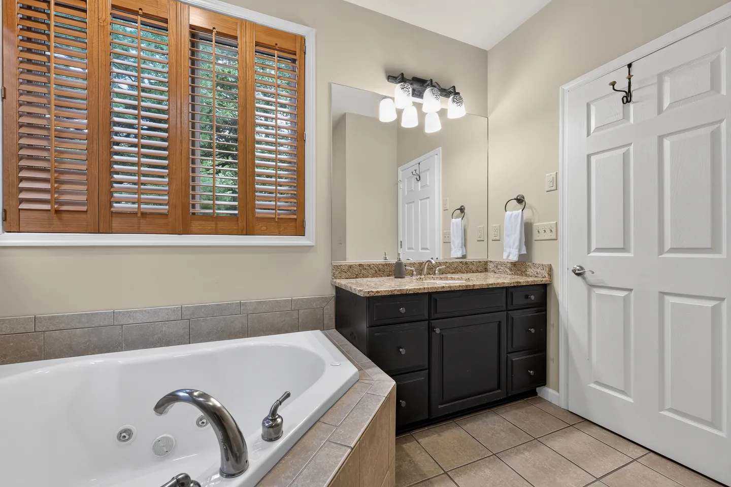 Bathroom featuring a deep soaking jetted tub, tiled floor, dark vanity with speckled countertop, and window with wooden shutters.