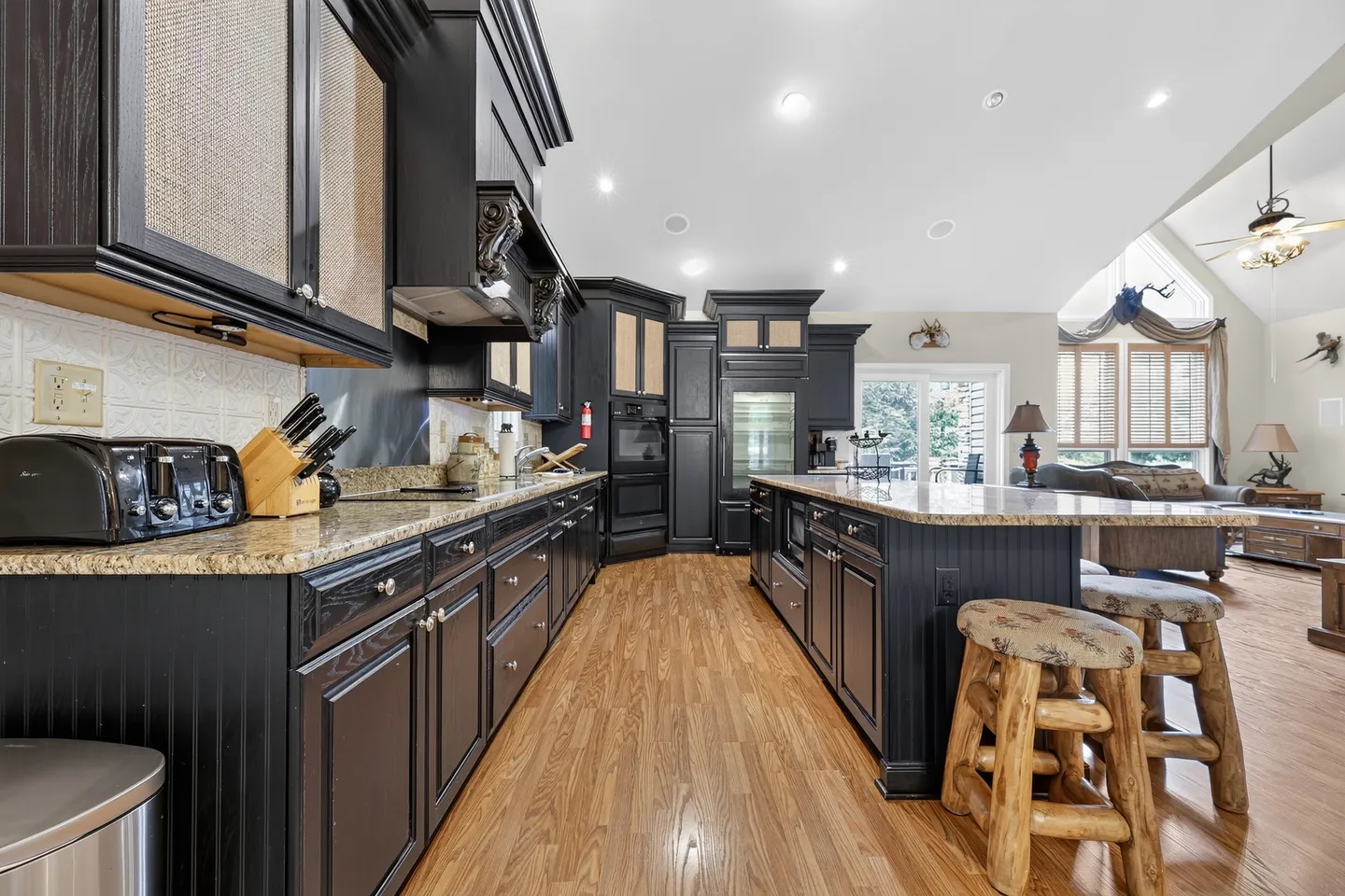 Spacious kitchen featuring dark cabinetry, speckled countertops, a large center island with log barstools, and wood-look flooring.