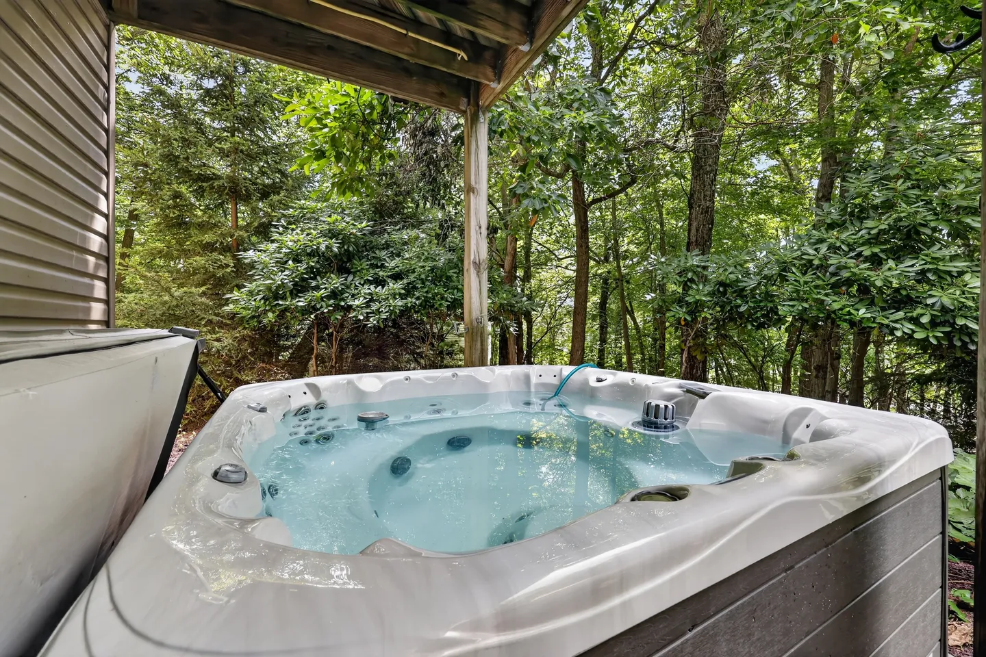 Outdoor hot tub filled with clear water situated beneath a covered wooden deck facing a dense green forest.
