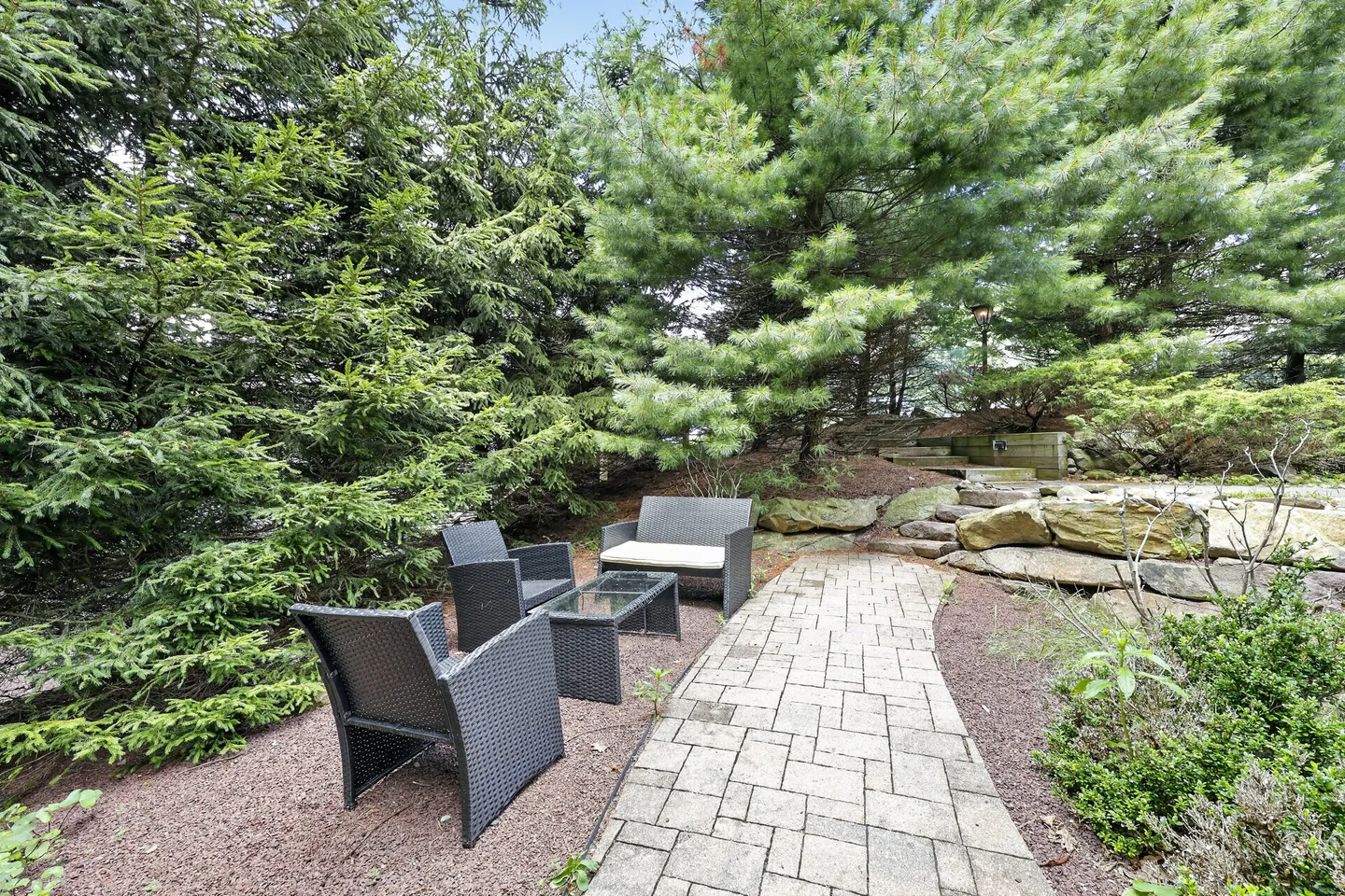 Outdoor paved walkway leading to an outdoor seating area with wicker armchairs, a bench, and a glass-top coffee table surrounded by evergreen trees.
