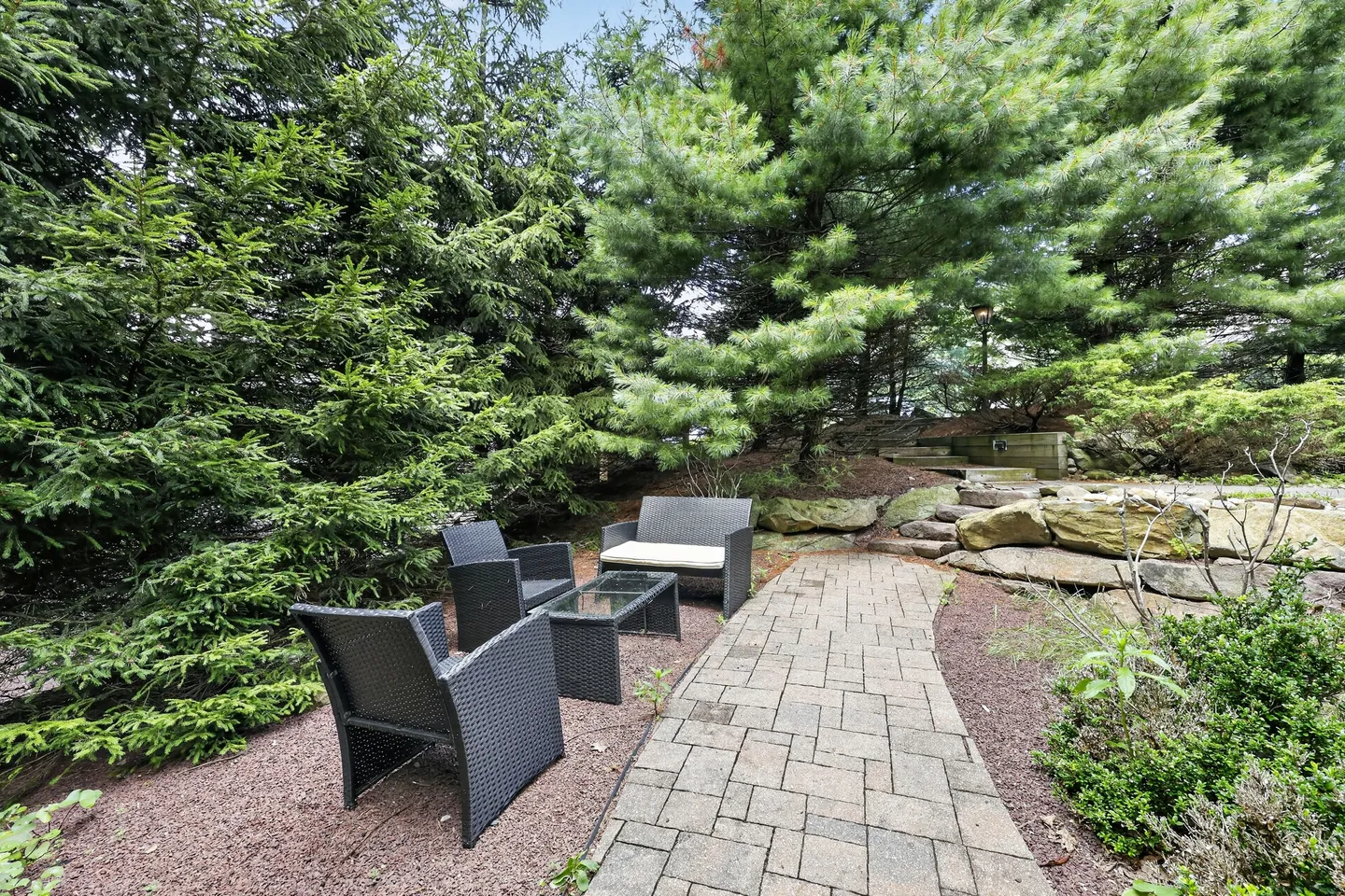 A stone paver pathway leads to outdoor wicker seating surrounded by evergreen trees and rock landscaping.