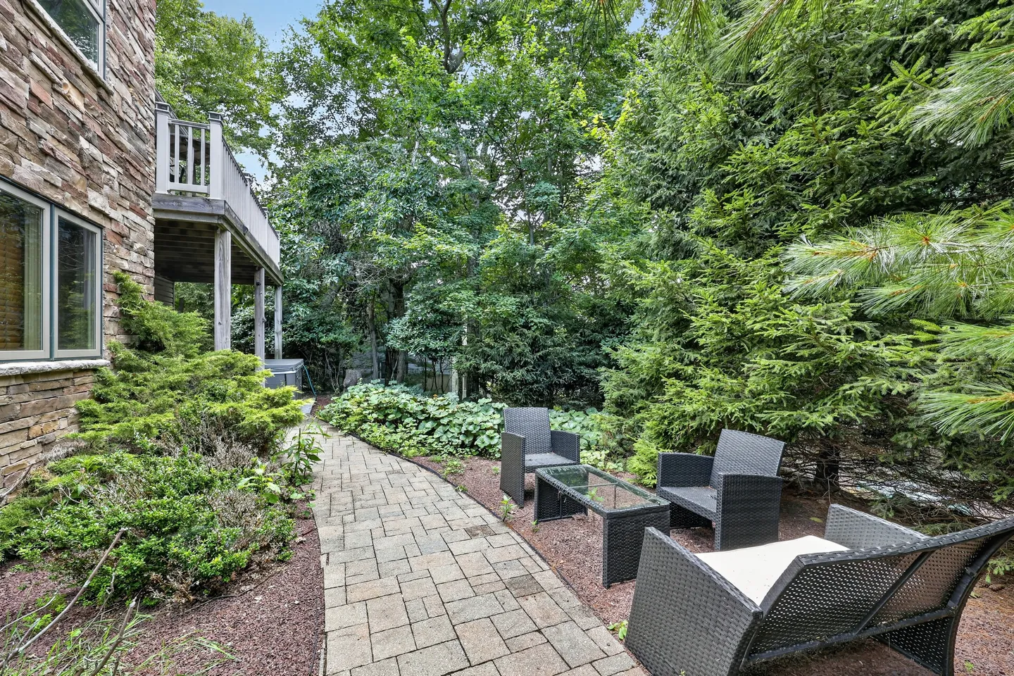 A stone walkway leading through a lush garden next to a stone building, with woven outdoor seating and a glass-topped coffee table.