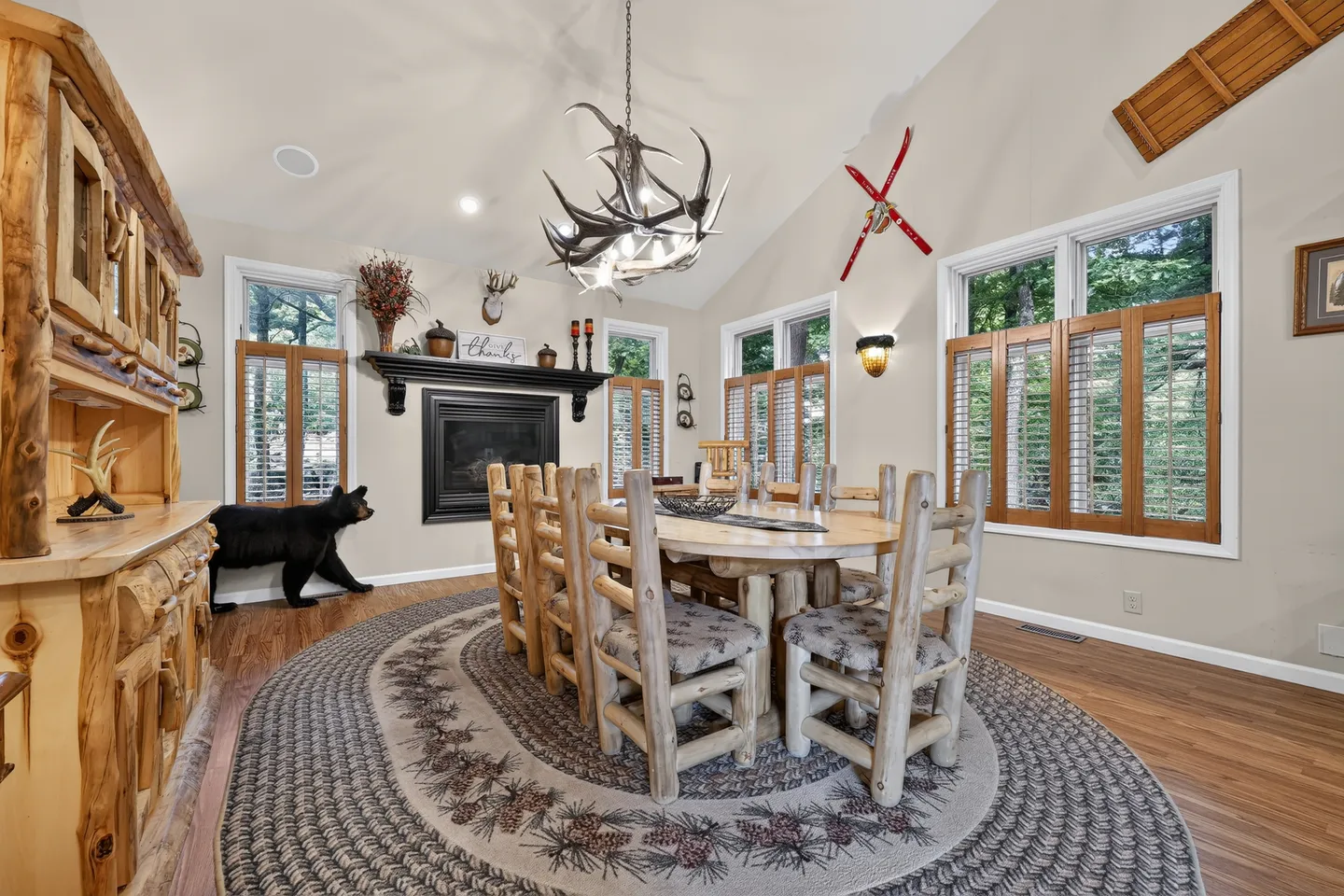 Rustic dining room featuring a log table and chairs set upon a large braided rug, with an antler chandelier, fireplace, and rustic decor.