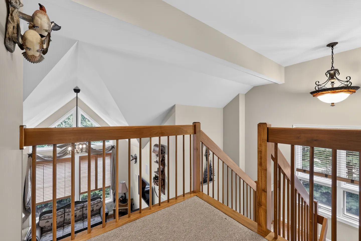View from an upper carpeted hallway overlooking a living area below, framed by wooden railings and showing vaulted white ceilings.